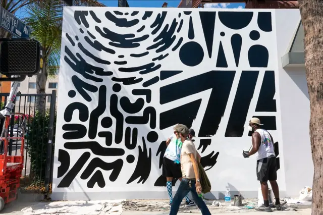 The Upendo wall going up on Martel Avenue, Los Angeles — the Open Air Gallery crew painting black shapes on a clean white wall, drop cloths on the pavement